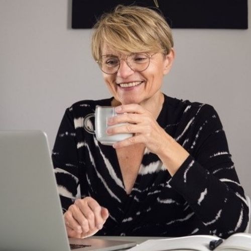 Eine Frau sitzt am Laptop, trinkt Kaffe und schaut dabei auf den geschriebenen Text.
